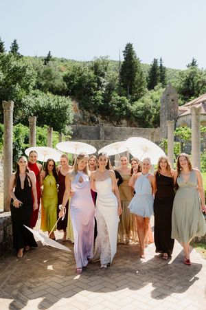 Bride in white gown walks with colorful bridesmaids holding parasols at an outdoor wedding venue.
