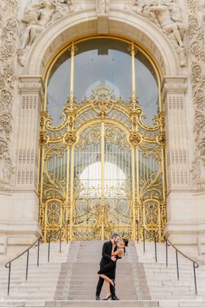 A couple embraces on grand marble steps in front of an ornate gold and white baroque gate in Paris.
