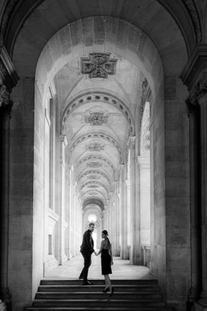 A silhouetted couple stands in a dramatic arched hallway with classical architecture and repeating vaulted ceilings.