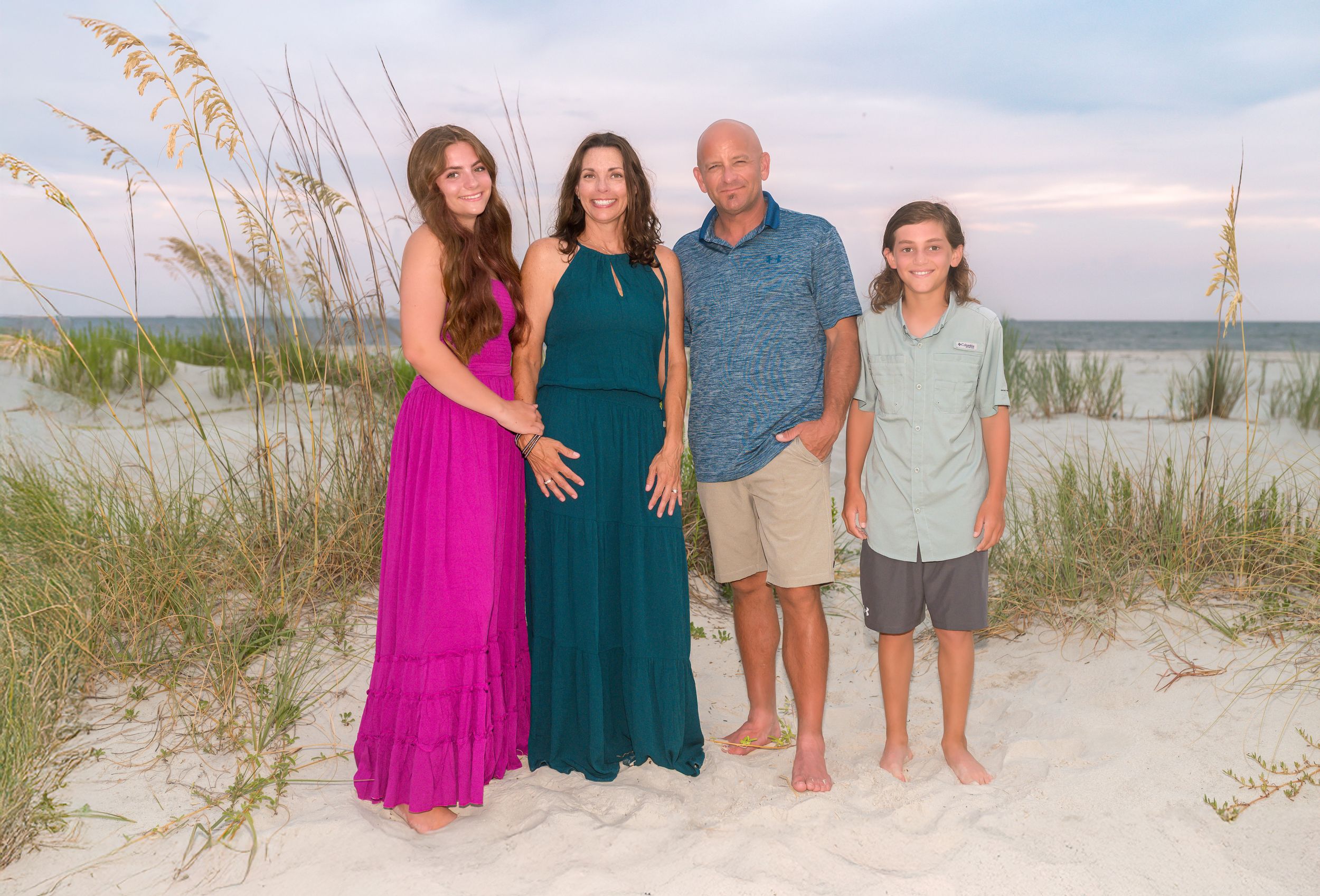 Family group poses together on sandy beach with sea oats and dunes during sunset photo session. Dauphin Island, Alabama