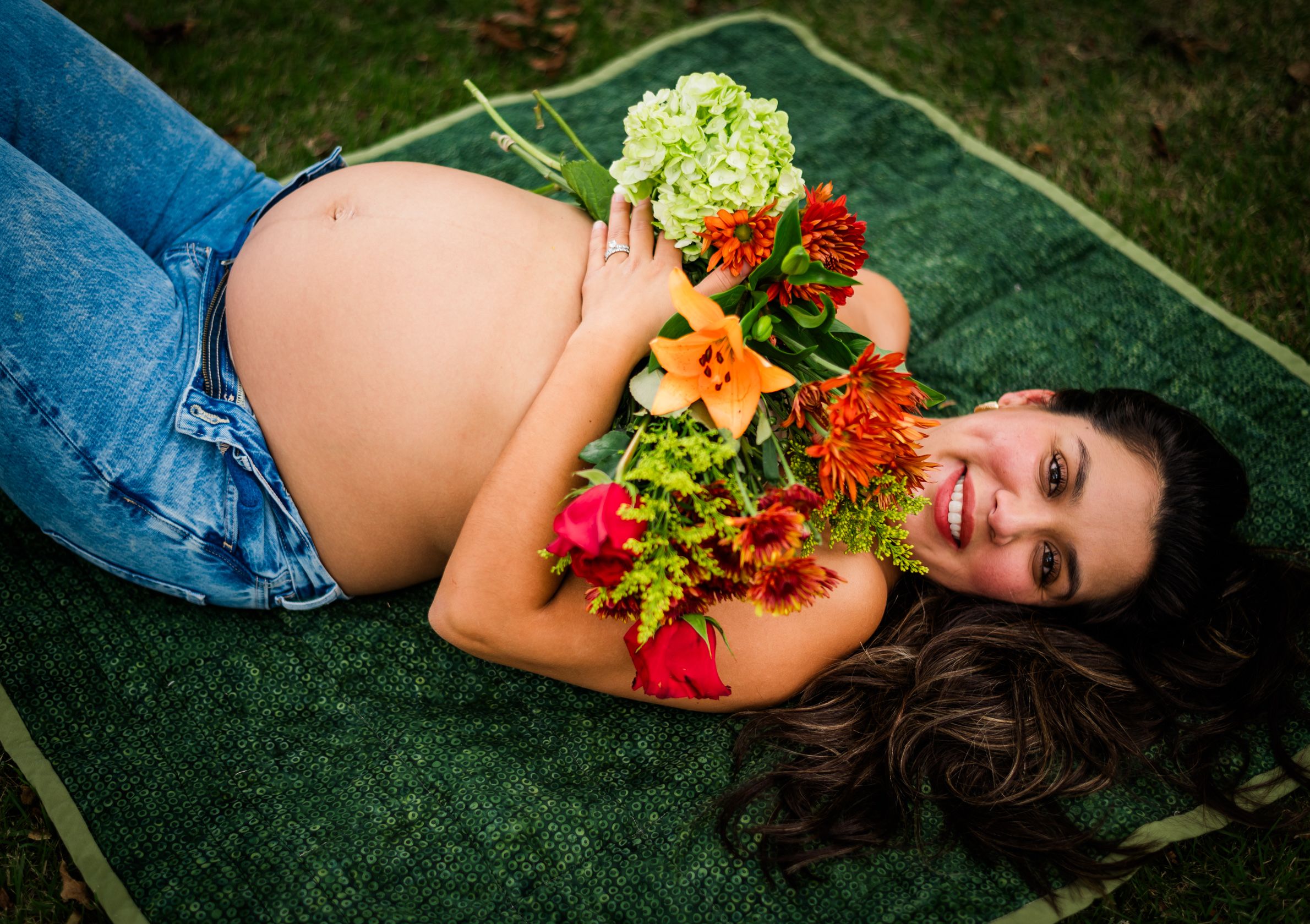 Woman in jeans lying on grass holding colorful flowers and vegetables in a maternity photo shoot.