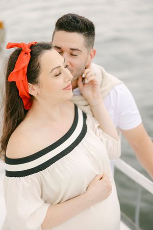 A couple shares a romantic moment on a boat, man kissing womans temple, she wears off-shoulder dress with red hair bow.