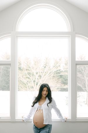 A pregnant woman in white sweater stands by bright sunlit windows during a maternity photoshoot.