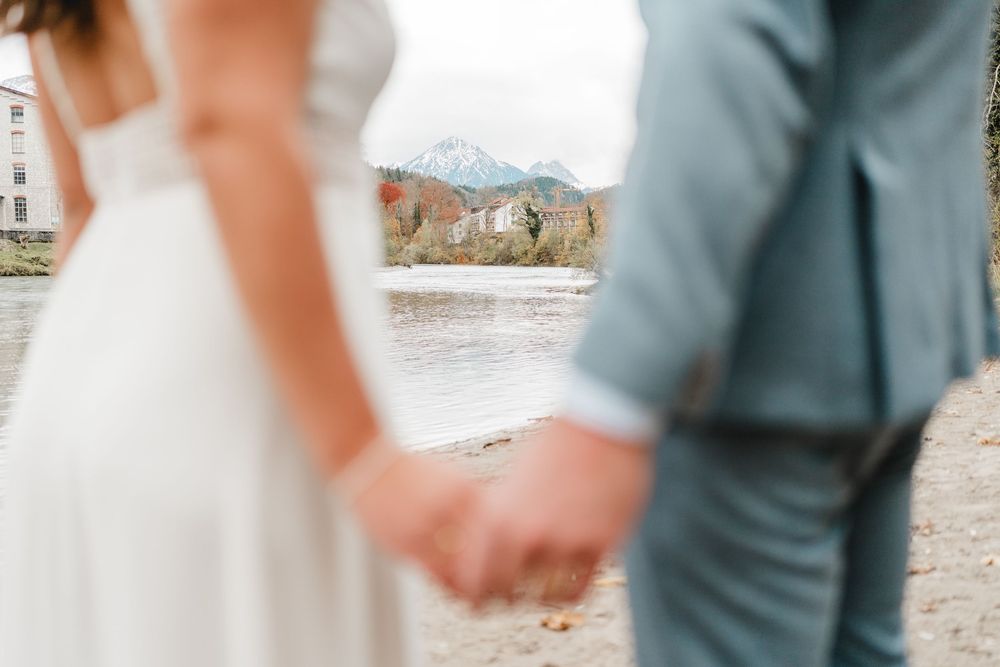 Detailbild von Händen eines Brautpaares mit dem Fokus der Berge. Fotografiert an der Lech in Füssen von Ramona Maier aus Schemmerhofen
