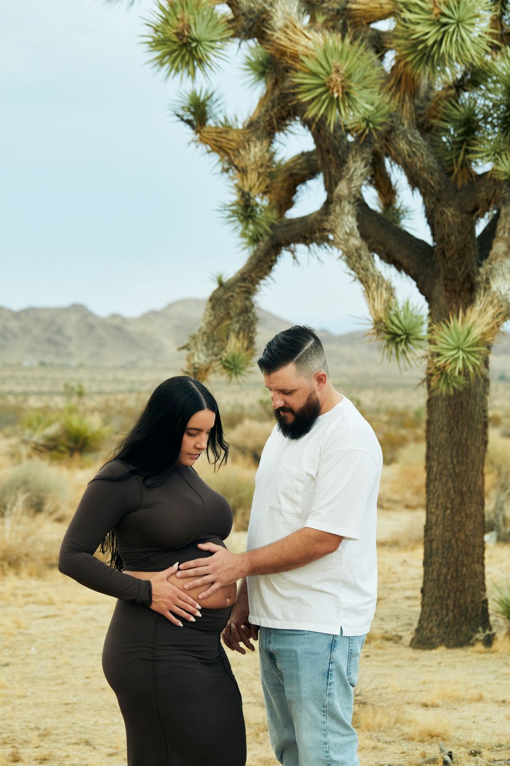 Couple sharing tender moment under Joshua tree in desert maternity photo shoot.