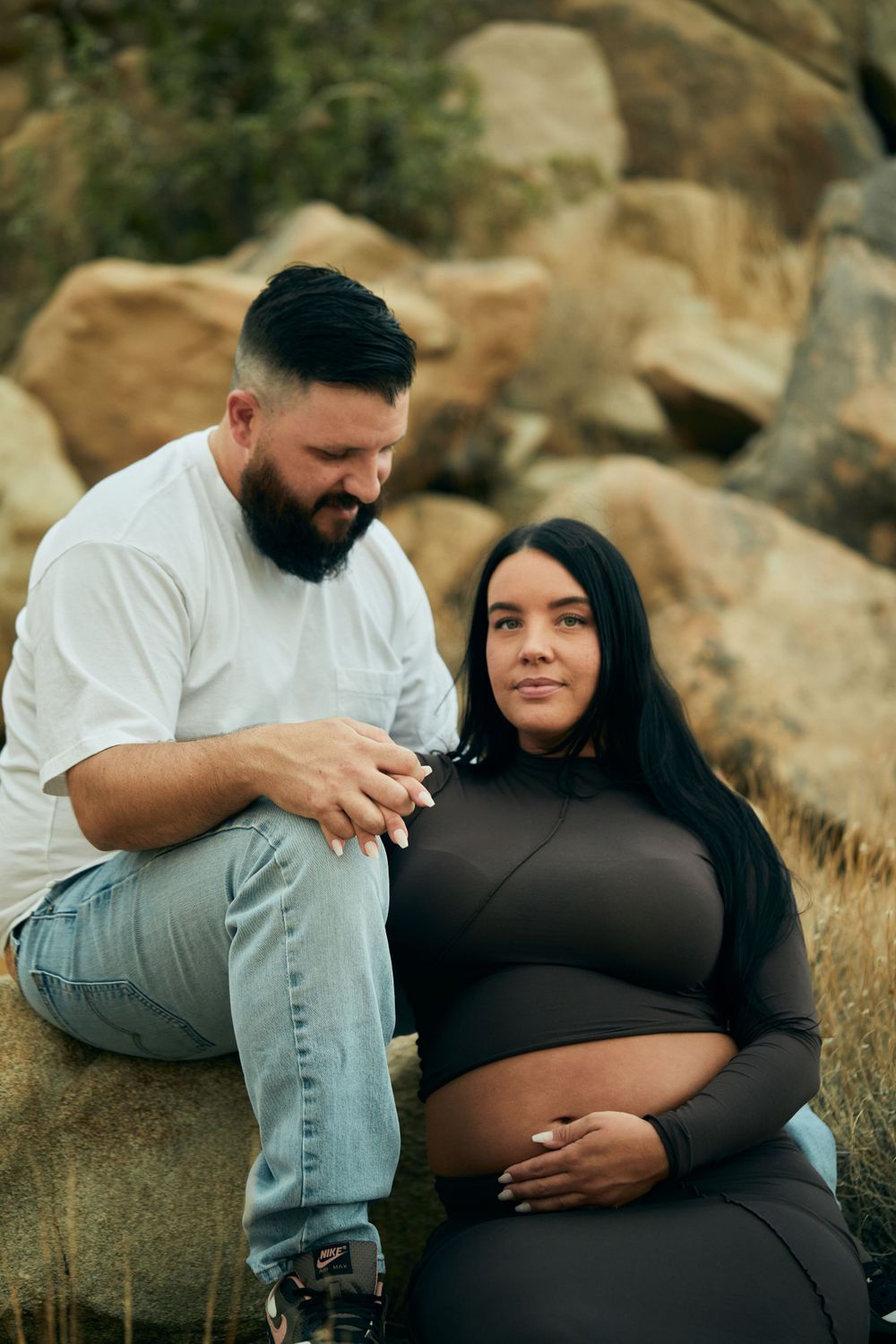 Expectant couple sitting on rocks share an intimate moment in an outdoor desert setting with rustic boulders.