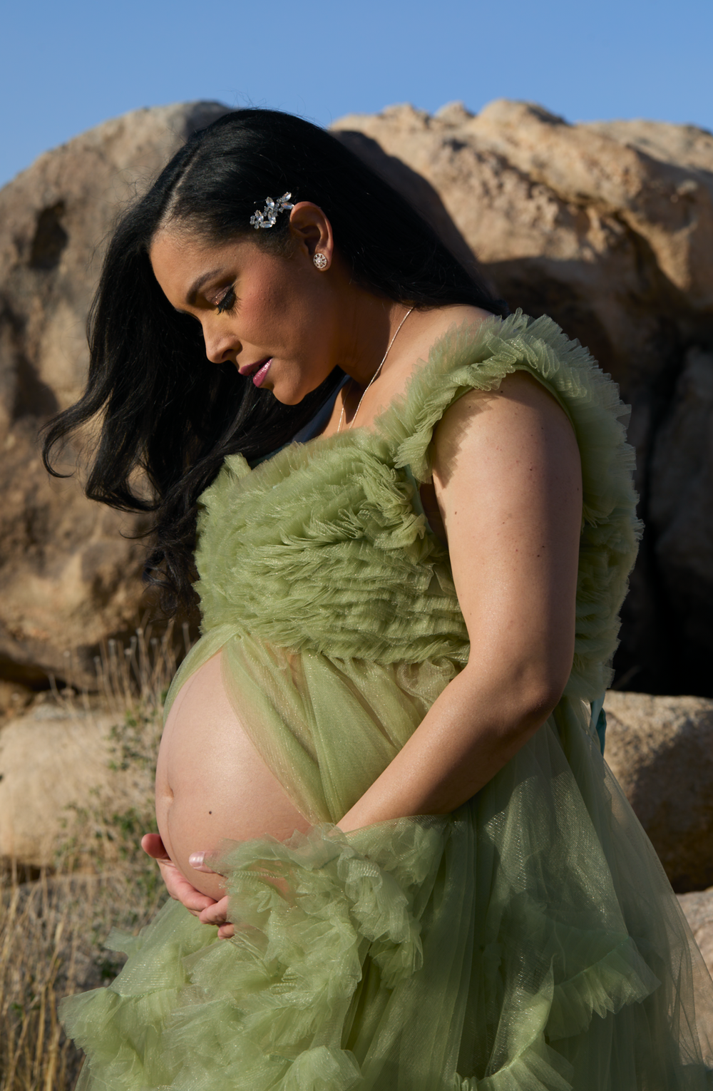 Expectant mother in flowing sage green dress poses against desert rock formations at sunset.