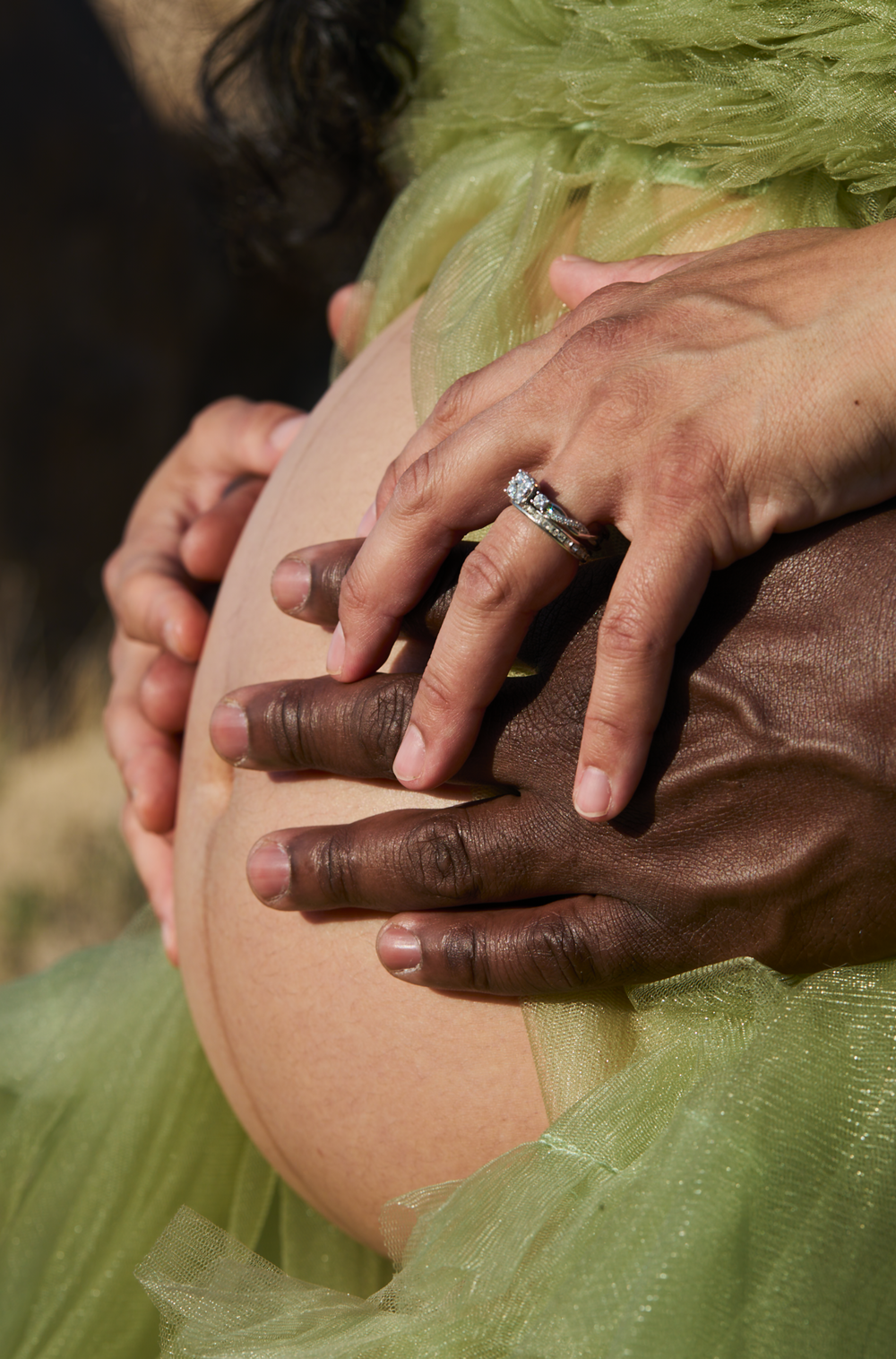 Intimate maternity photo showing hands cradling a pregnant belly through green tulle fabric with a diamond ring.