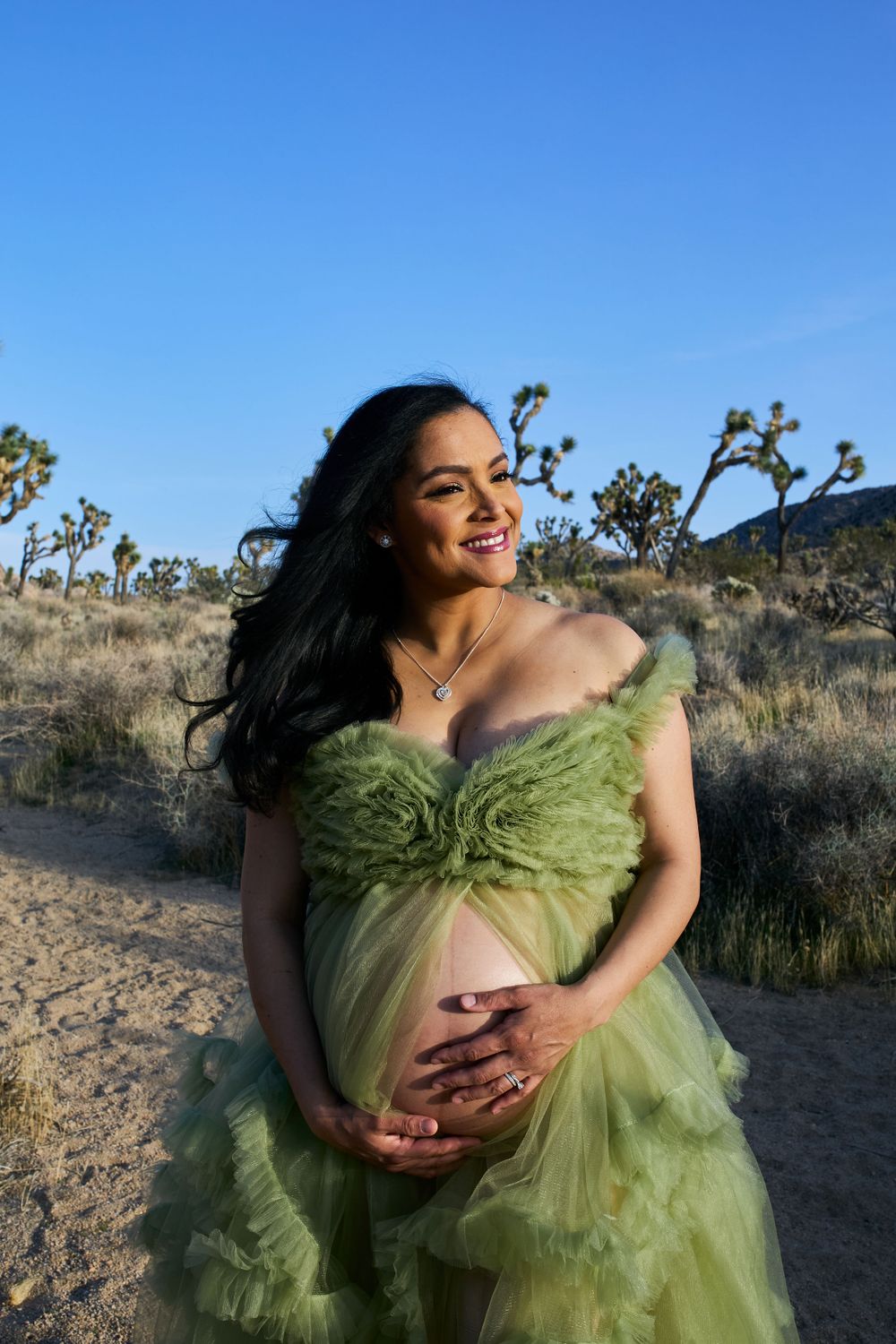An expectant mother in a flowing green dress poses in a desert setting with Joshua trees.