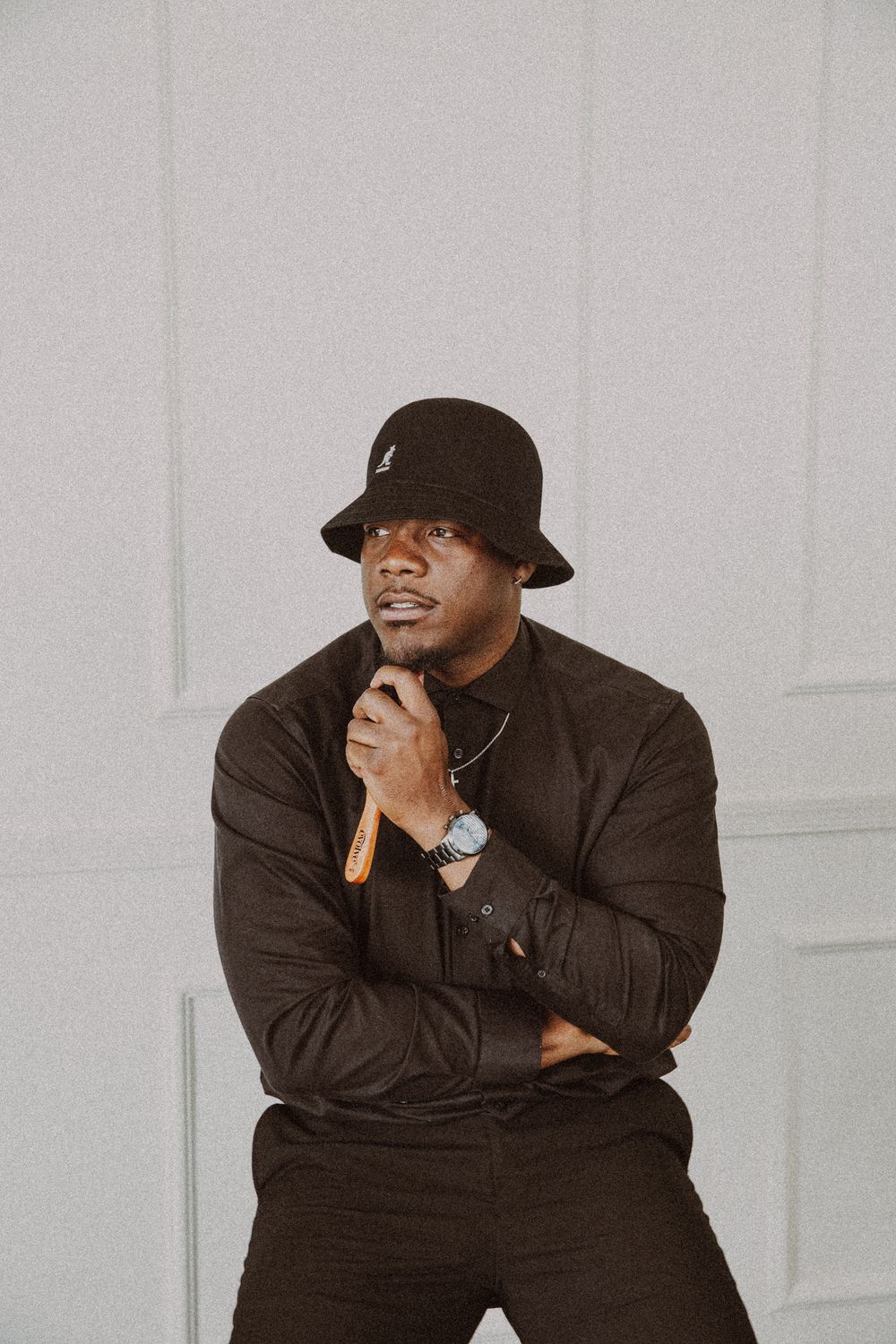 Confident Black man in black outfit and bucket hat posing thoughtfully against white wall.