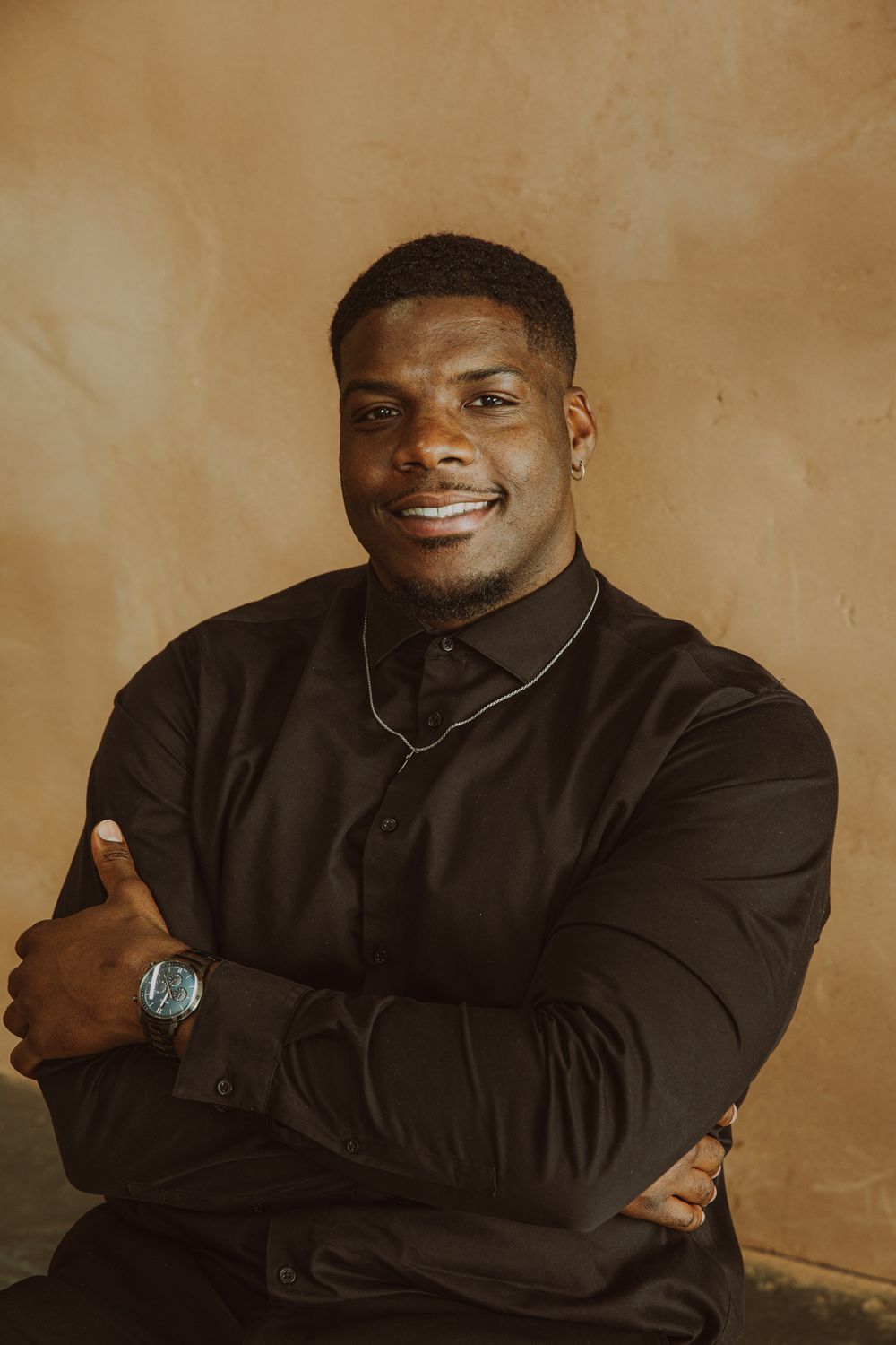 Smiling Black man in black shirt with watch and necklace posing arms crossed against tan wall.