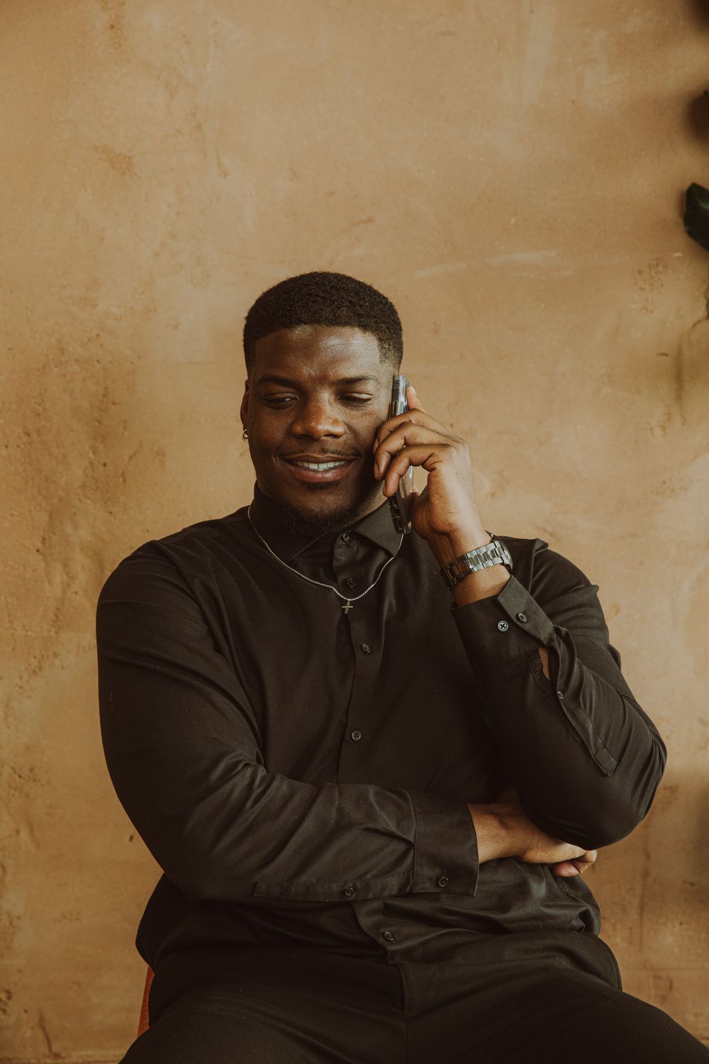 Smiling Black man in black shirt talking on phone against warm tan textured background.