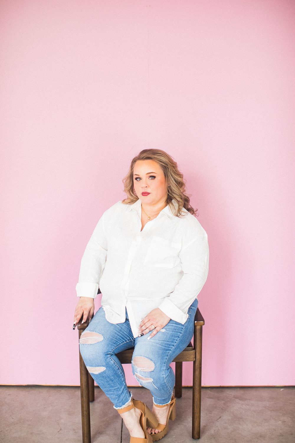 Plus-size woman in white shirt and ripped jeans sitting on chair against pink background.