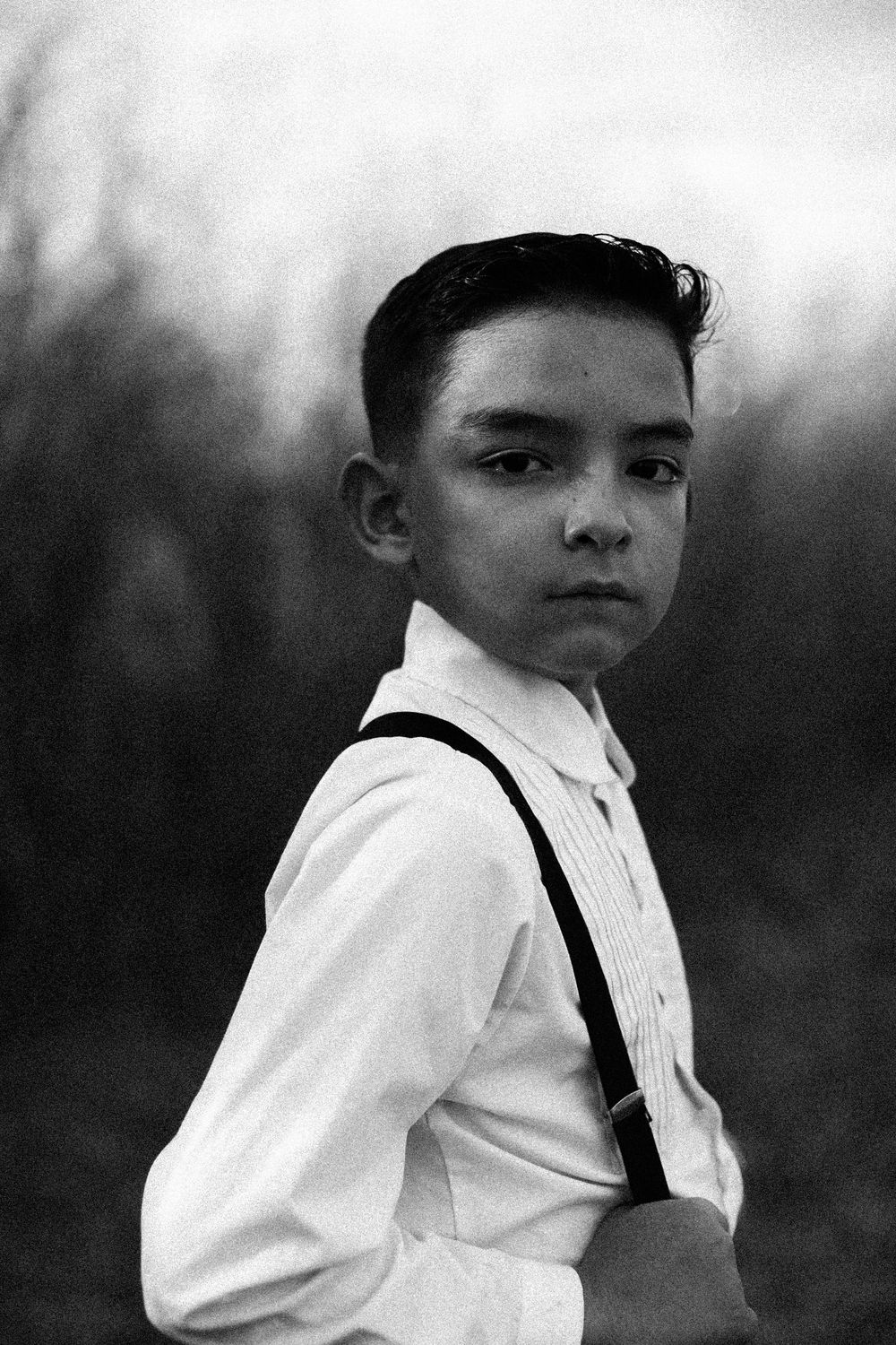 Black and white portrait of a young boy in white shirt with suspenders looking over shoulder outdoors.