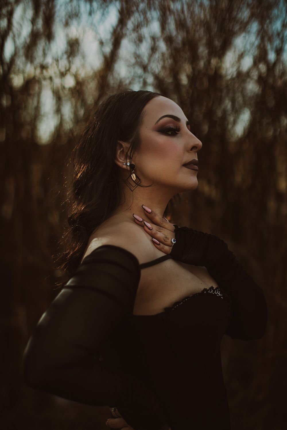 Woman in black off-shoulder dress poses outdoors at dusk, hand on shoulder, wearing statement earrings and bold makeup.