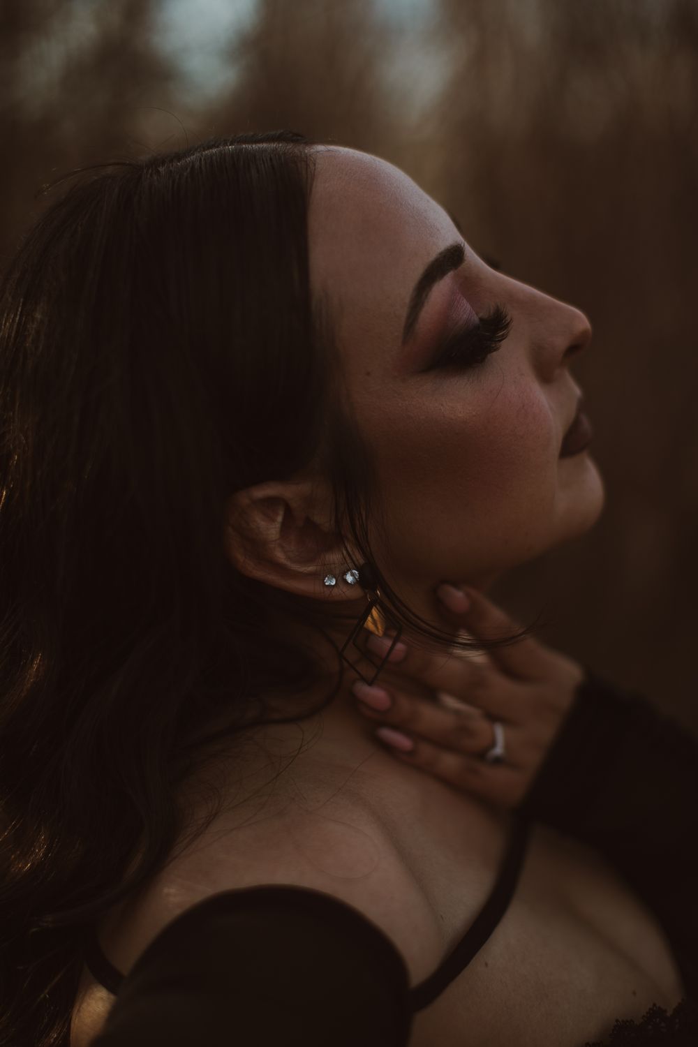 Close-up side profile of a woman with dramatic makeup, hand on neck, wearing earrings in moody outdoor lighting.