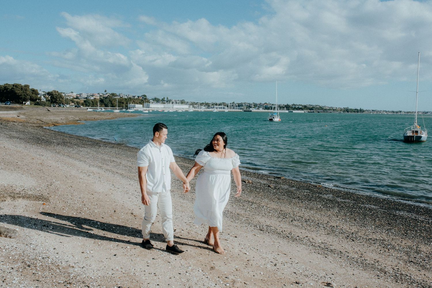 Happy couple in white outfits walking hand in hand along a sunny beach with sailboats in the distance.