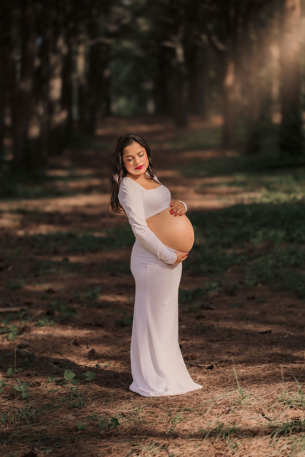 Expectant mother in white dress poses elegantly on wooded path during maternity photoshoot.