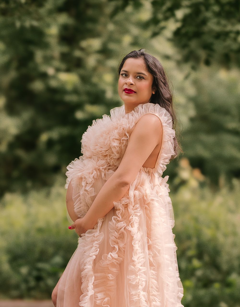 Pregnant woman in peach lace dress poses in outdoor woodland setting.