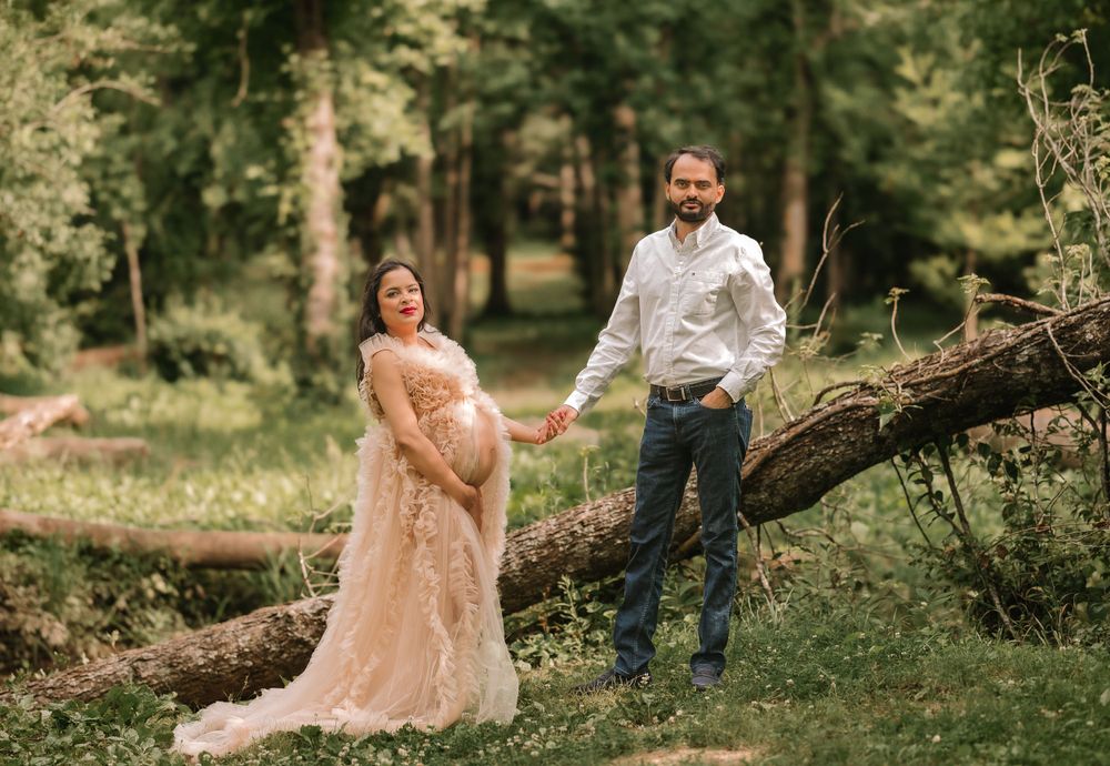 Maternity photoshoot in forest with couple posing near fallen tree in soft pink dress.