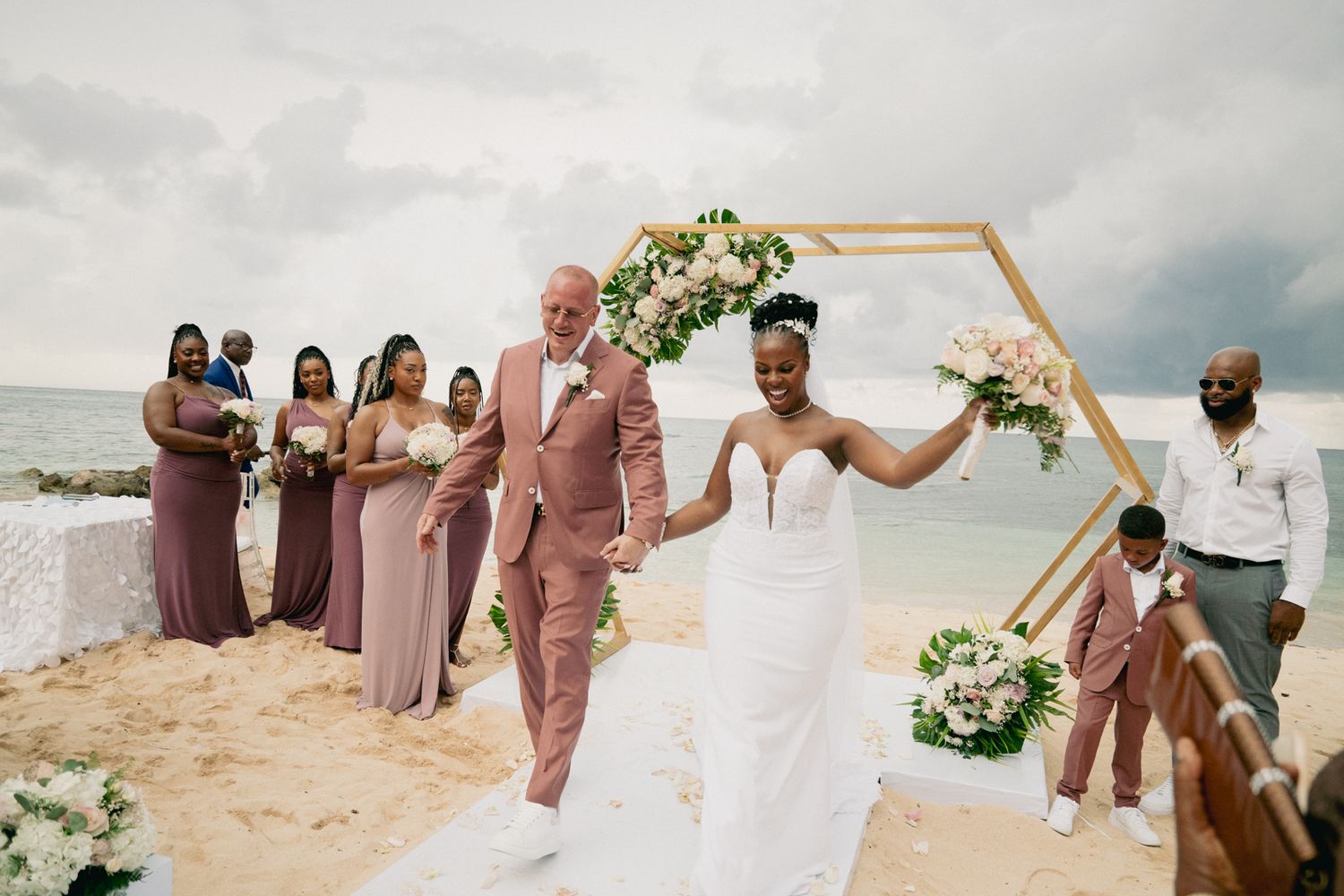 A beach wedding ceremony under a hexagonal arch decorated with white flowers on a sandy shore by the ocean.