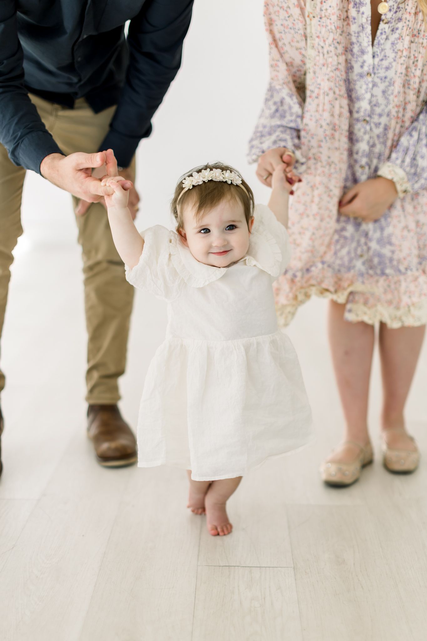 A baby in a white dress and floral headband takes first steps while being supported by parents on a light wood floor.