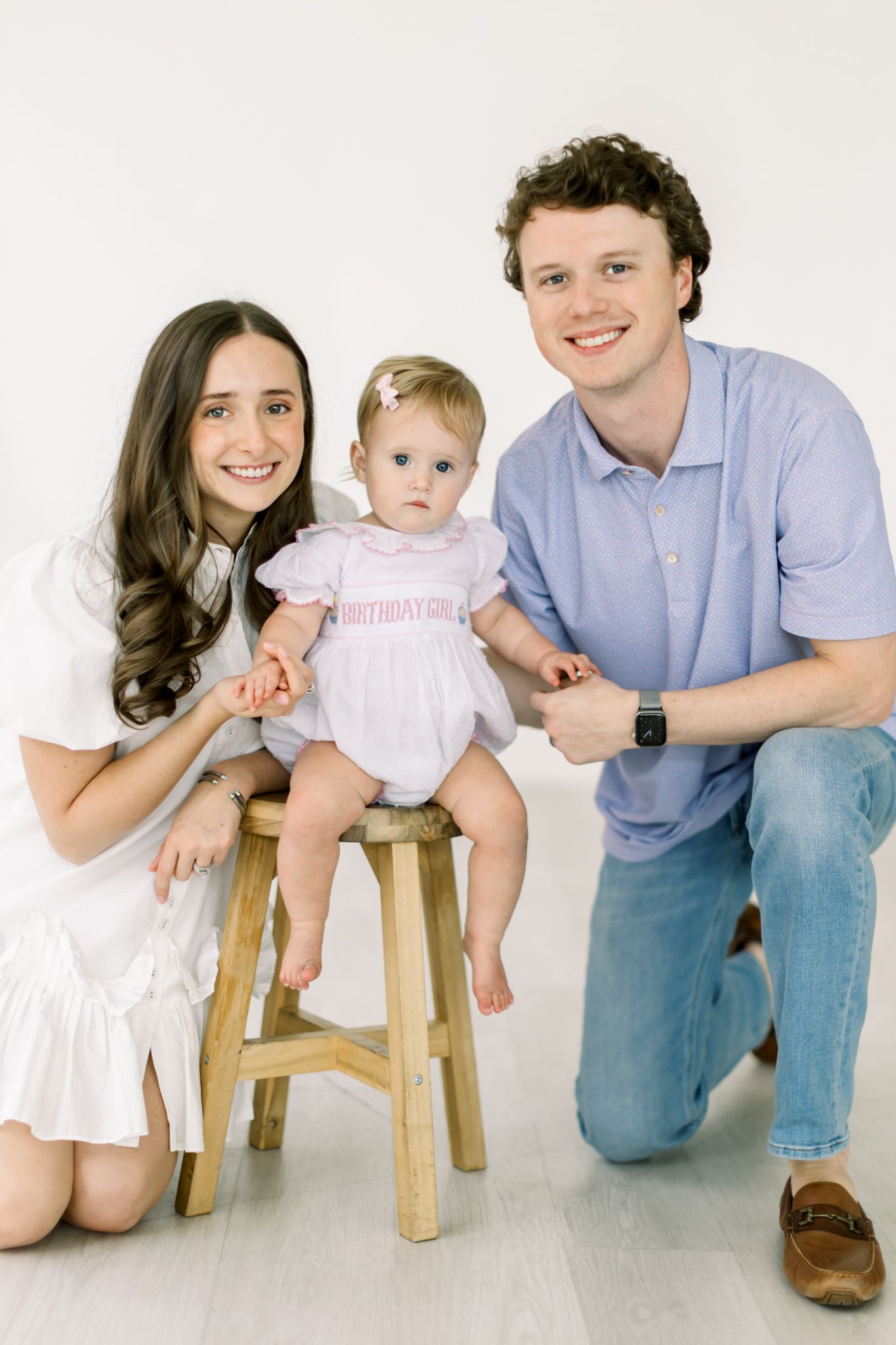 A family portrait with parents and baby on a wooden stool against a light background.