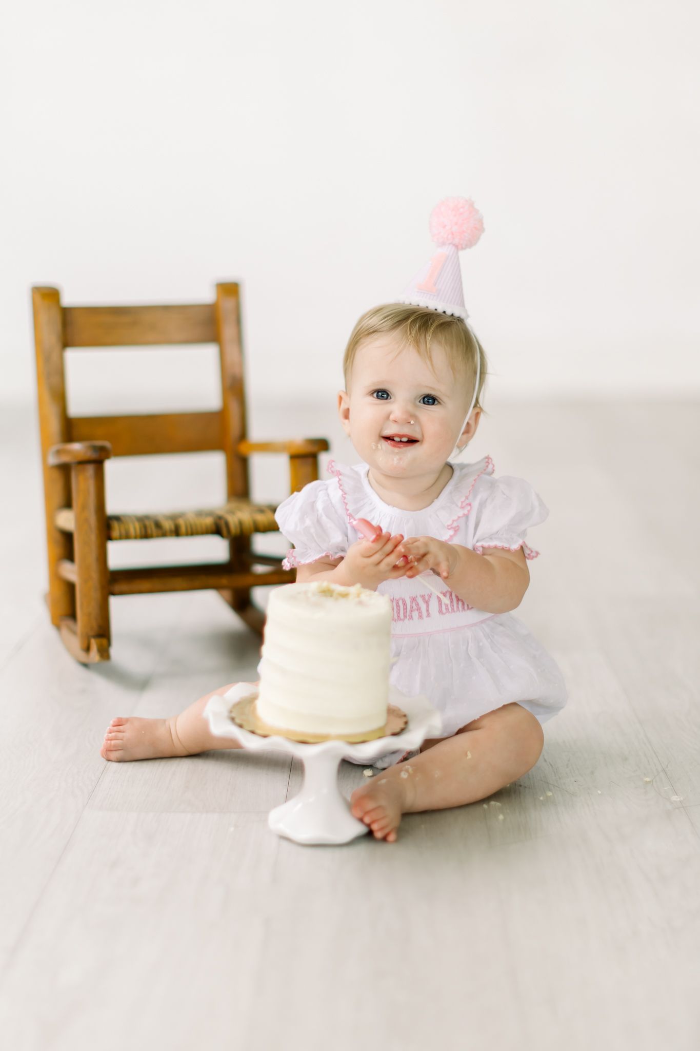 Sweet first birthday celebration scene with white cake on pedestal, pink party hat and wooden rocking chair in background.