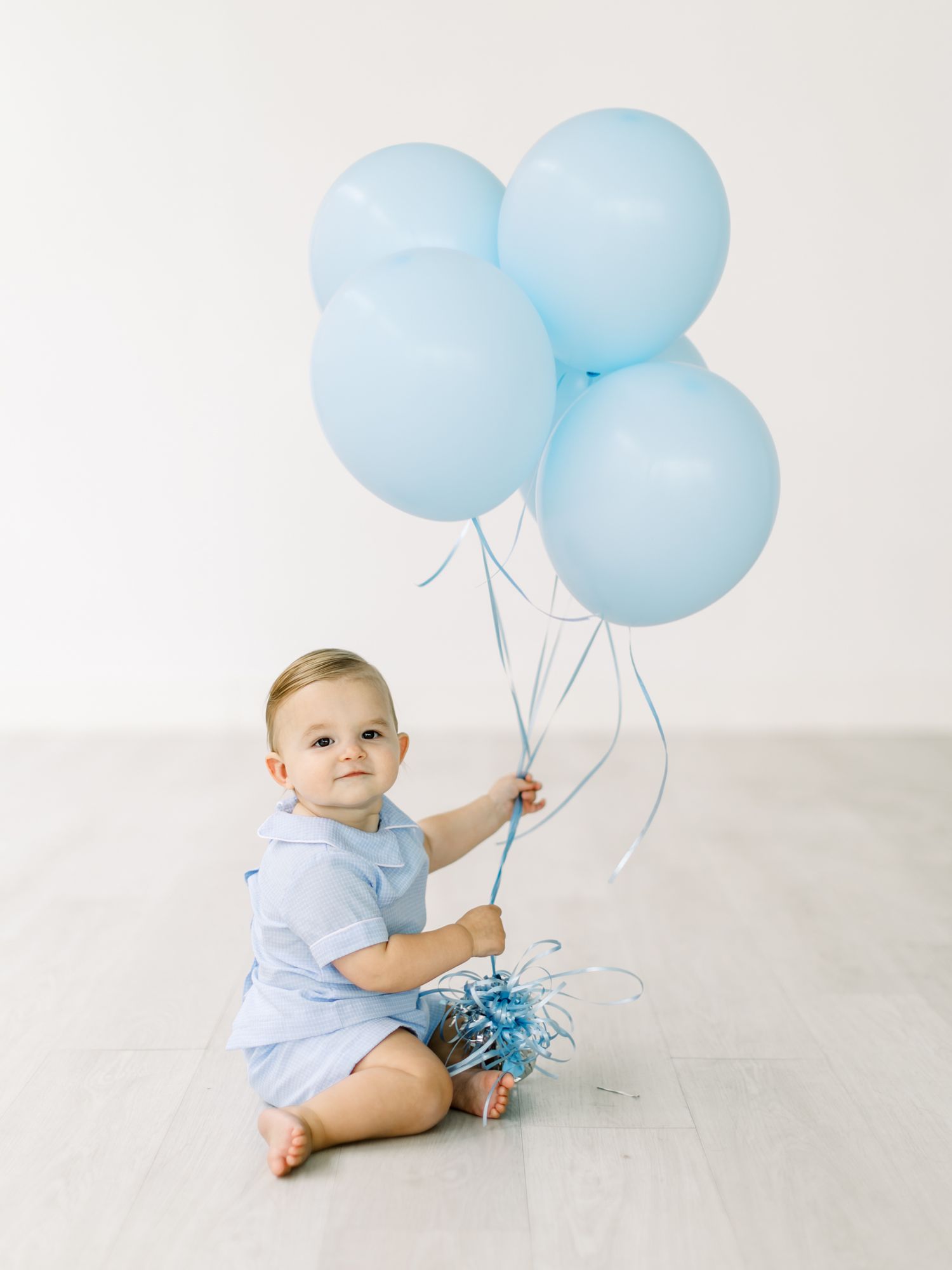 Small child in light blue outfit sits on white floor holding four pastel blue balloons during a photo session.