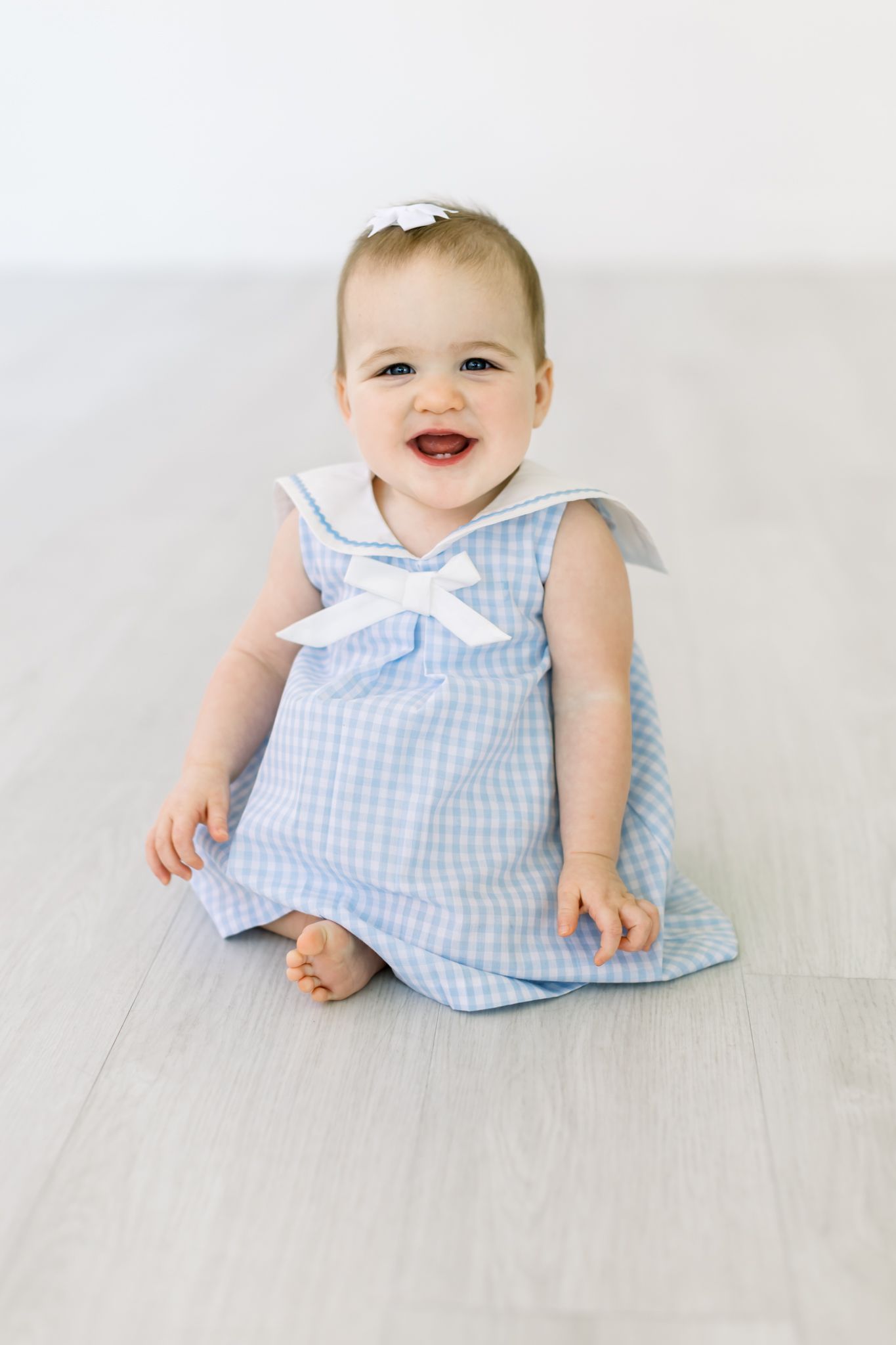 A baby wearing a light blue gingham dress with a bow sits smiling on a light wooden floor.