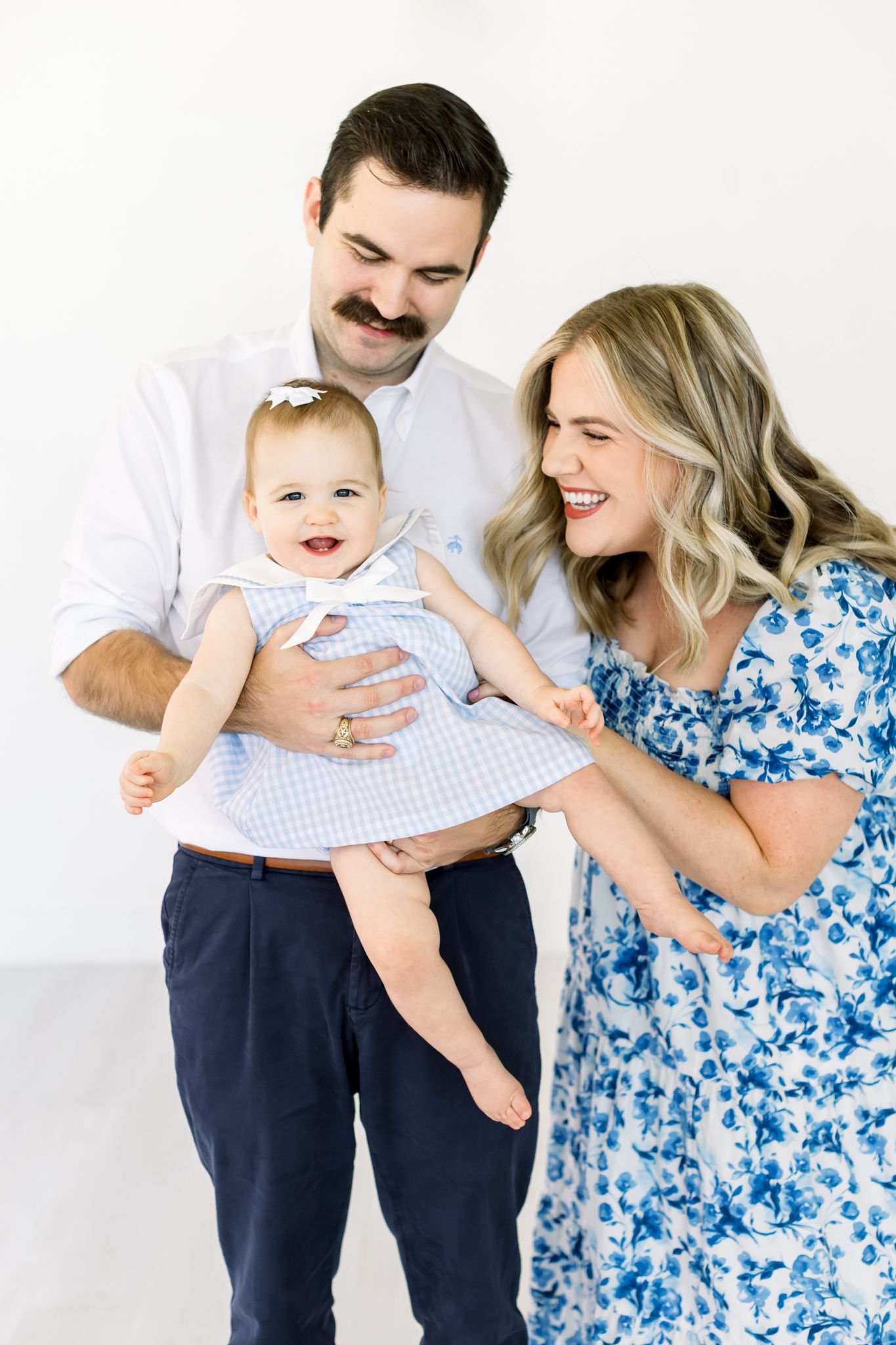 A happy family portrait with an infant in a gingham dress against a white background.