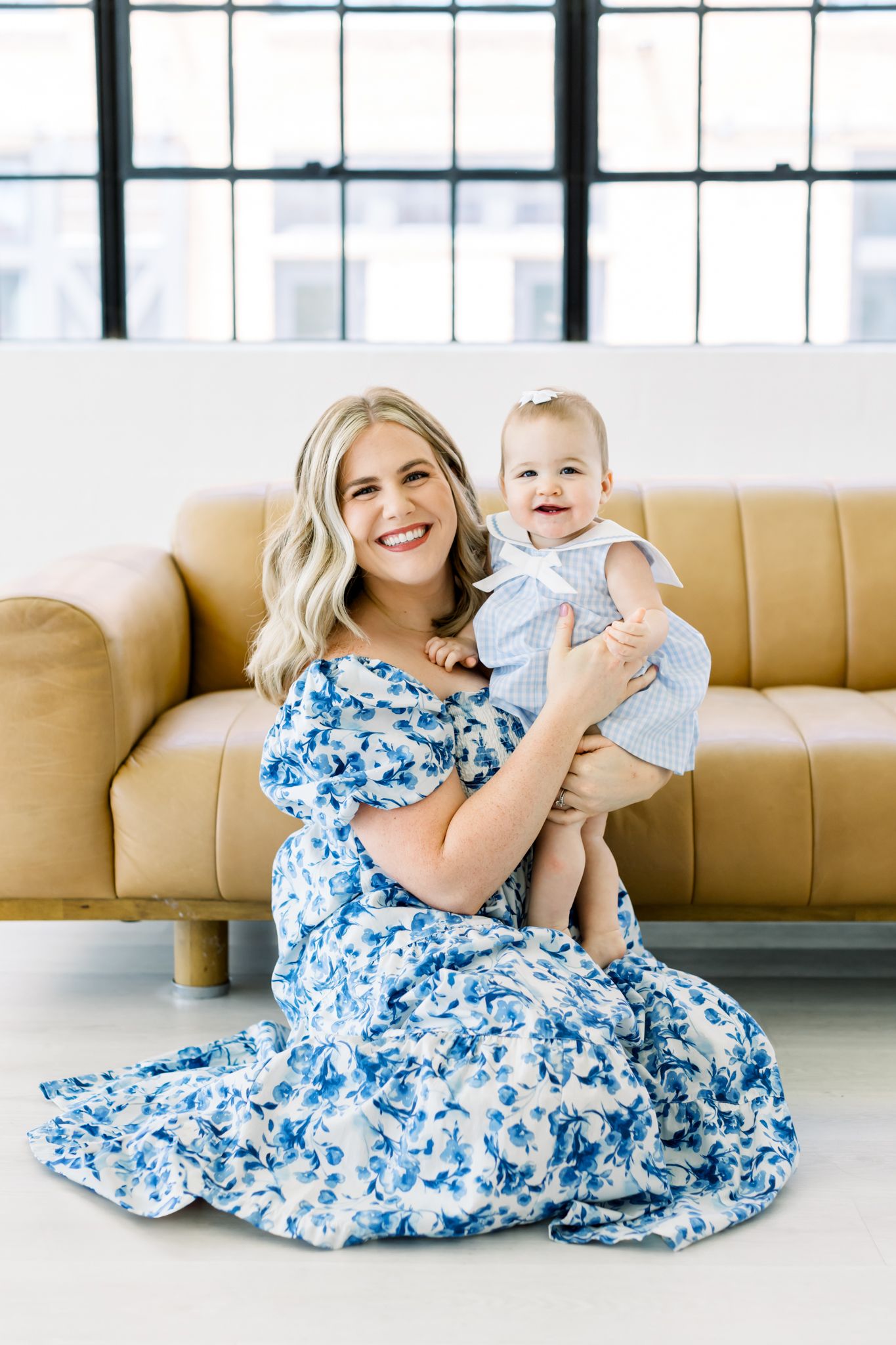 A woman in a blue and white floral dress sits on the floor with a baby in light blue clothing beside a tan leather couch.