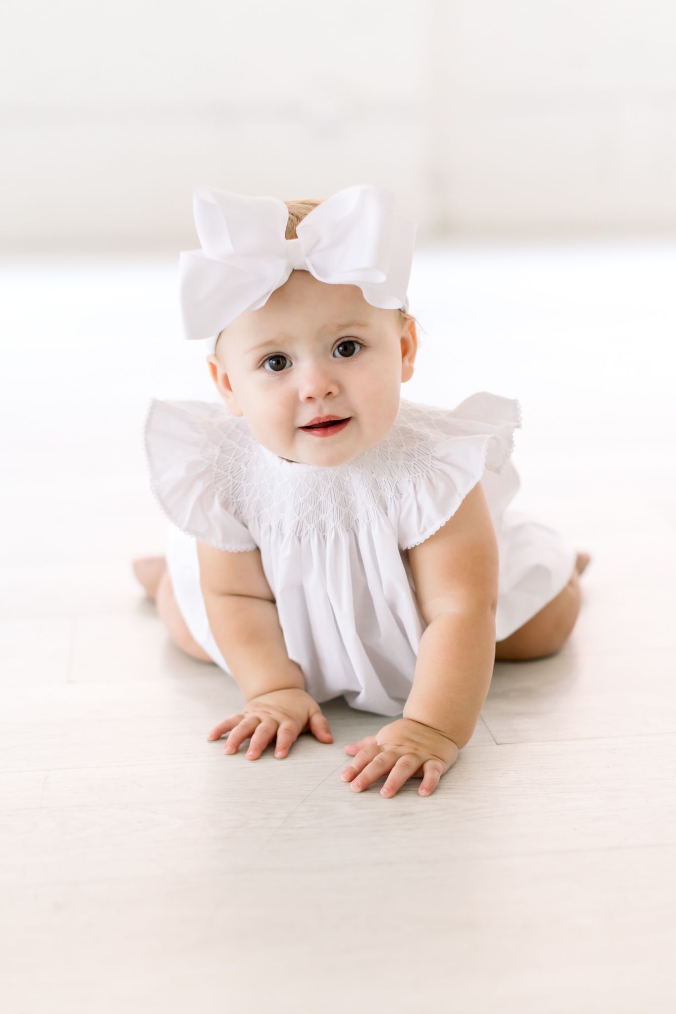 A baby in a white dress with ruffles and a bow headband crawls on a light floor.