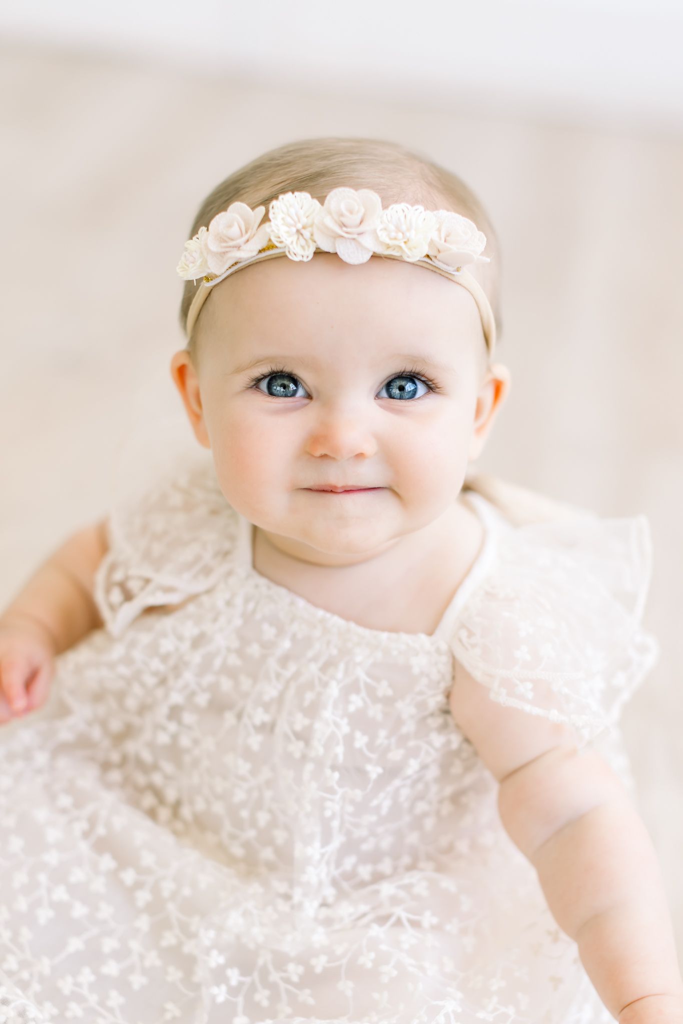 A baby wearing a delicate white lace dress and floral headband smiles at the camera with bright blue eyes.