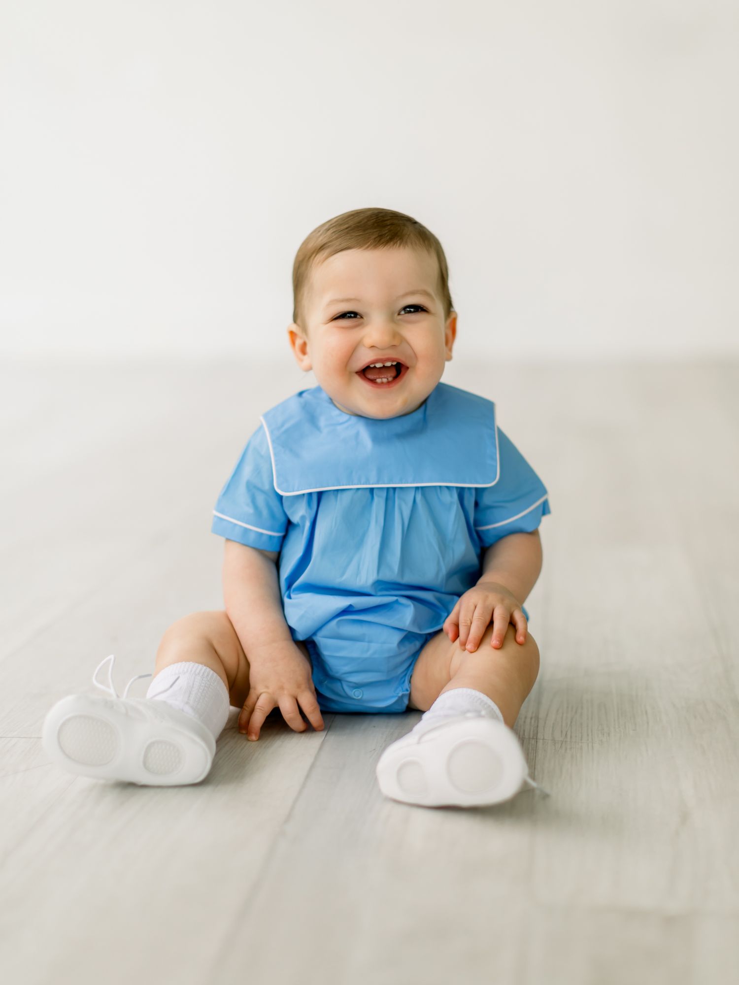 Adorable baby in light blue romper outfit and white shoes sits smiling on light wooden floor.