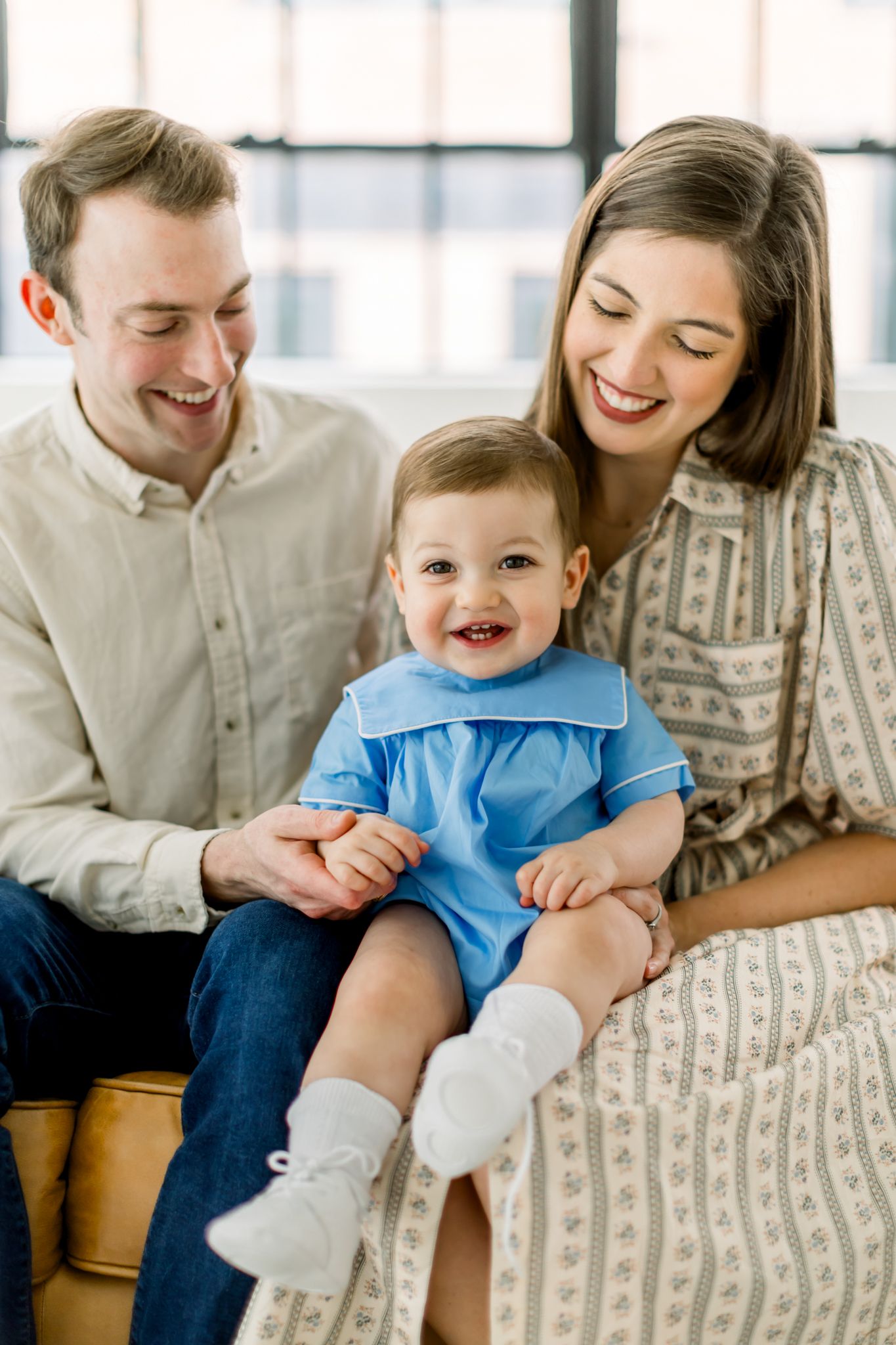 Young family sits together on a sofa with a laughing toddler in a light blue shirt.