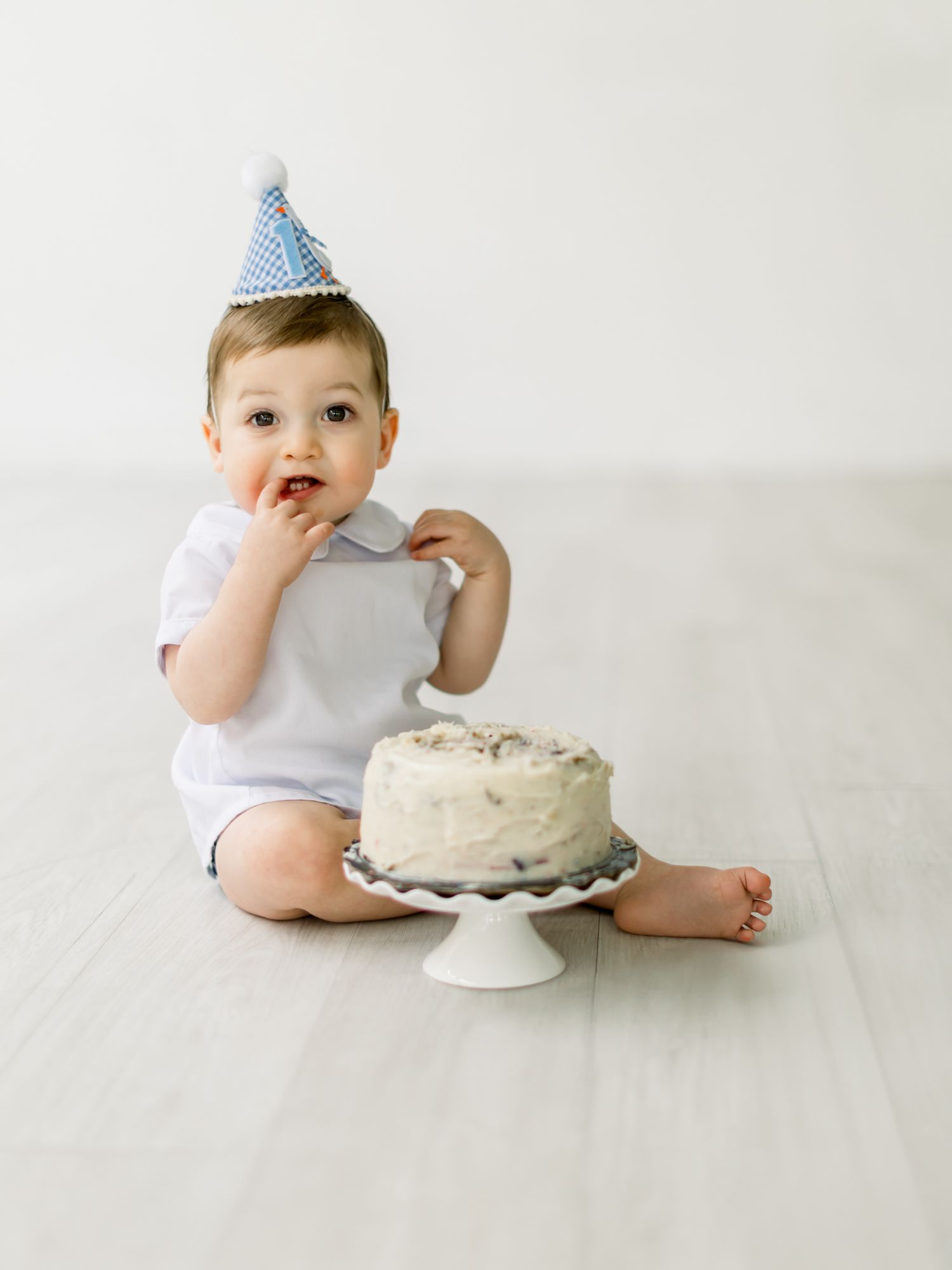 Small toddler in blue party hat sits on wooden floor with white birthday cake on cake stand beside them.