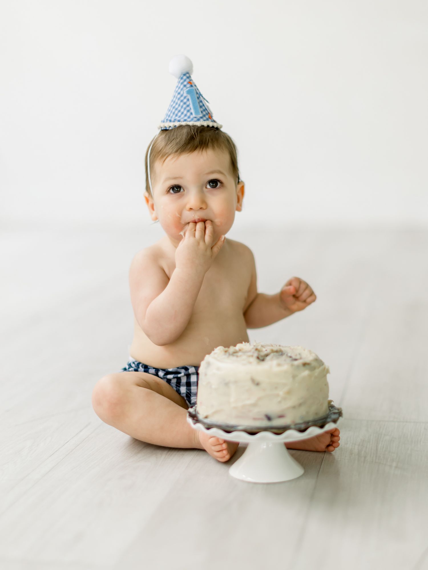 Baby wearing blue party hat and checkered diaper sits next to white birthday cake on cake stand against white backdrop.