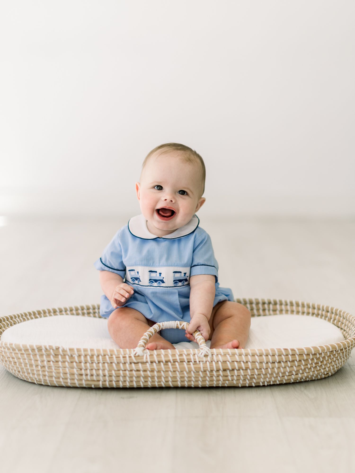 Baby wearing light blue romper sits happily in a woven changing basket against white background.