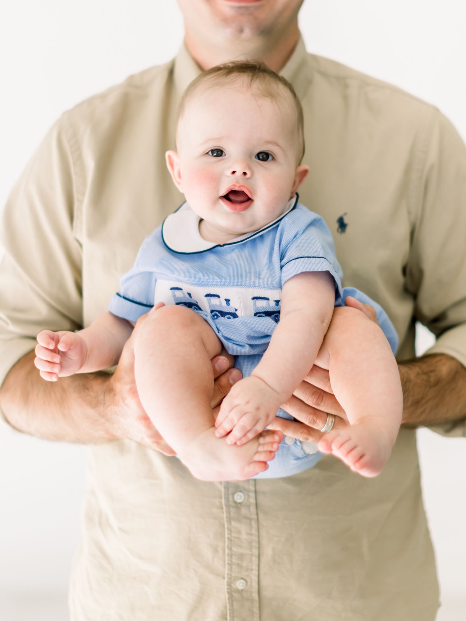 A baby in a light blue outfit sits contentedly on someone wearing a beige button-down shirt.