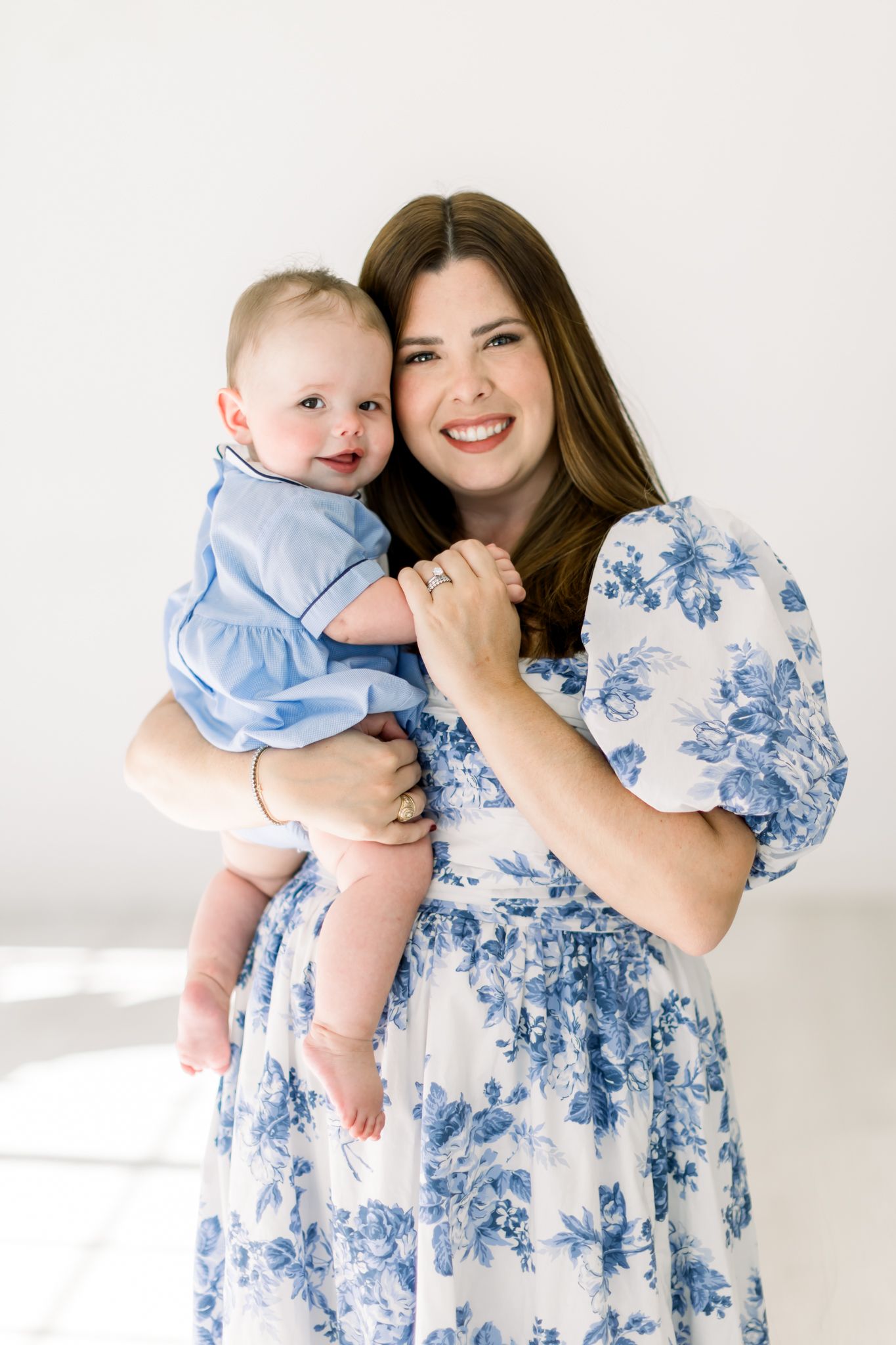 A joyful portrait showing a baby in light blue clothing and an adult wearing a floral blue and white dress against a white backdrop.