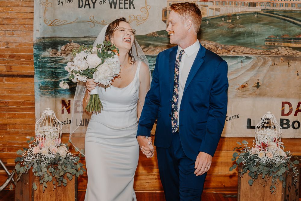 Joyful wedding couple laughing together in front of rustic wooden backdrop with floral arrangements.