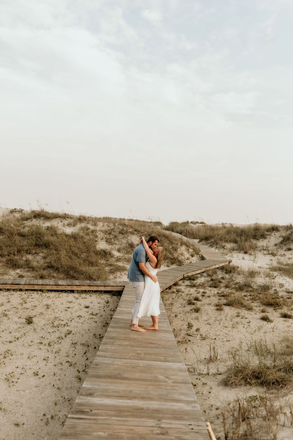 Couple embracing on a wooden boardwalk path through sandy beach dunes under a soft cloudy sky.