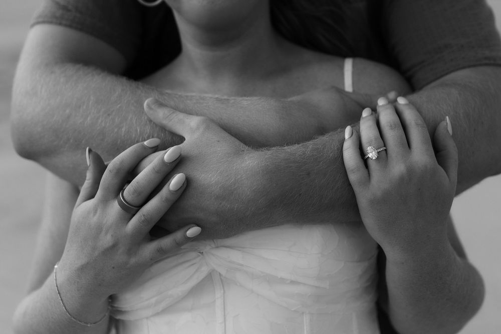 Black and white photo of a couple embracing, showing their hands and wedding rings in a romantic pose.