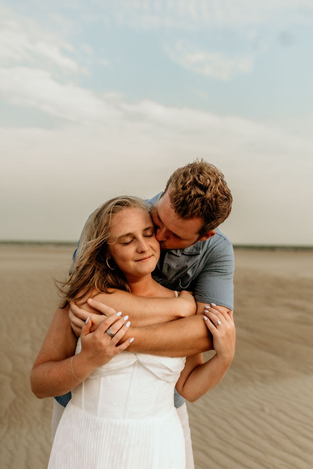A couple embraces tenderly on a sandy beach, the man hugging the woman from behind as she smiles with eyes closed.