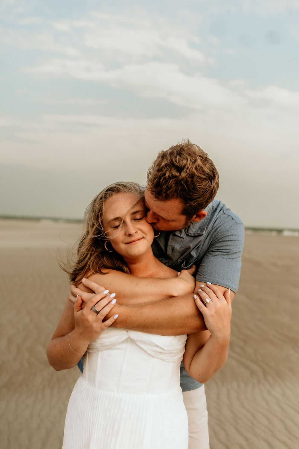 Couple embracing on a sandy beach, woman in white dress with eyes closed, man hugging her from behind under cloudy sky.