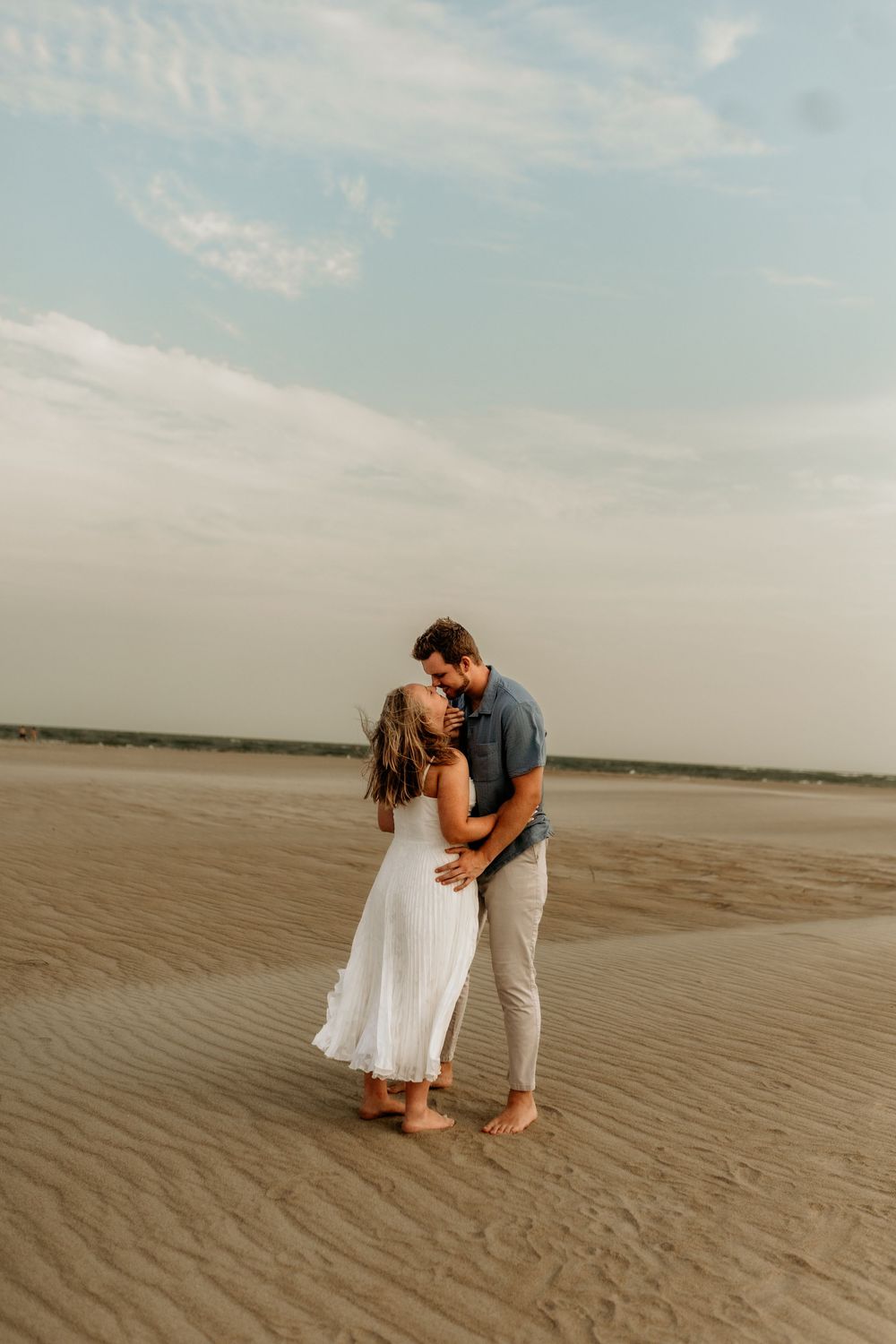 Couple sharing a romantic kiss on a sandy beach, woman in white dress, man in blue shirt and khaki pants.