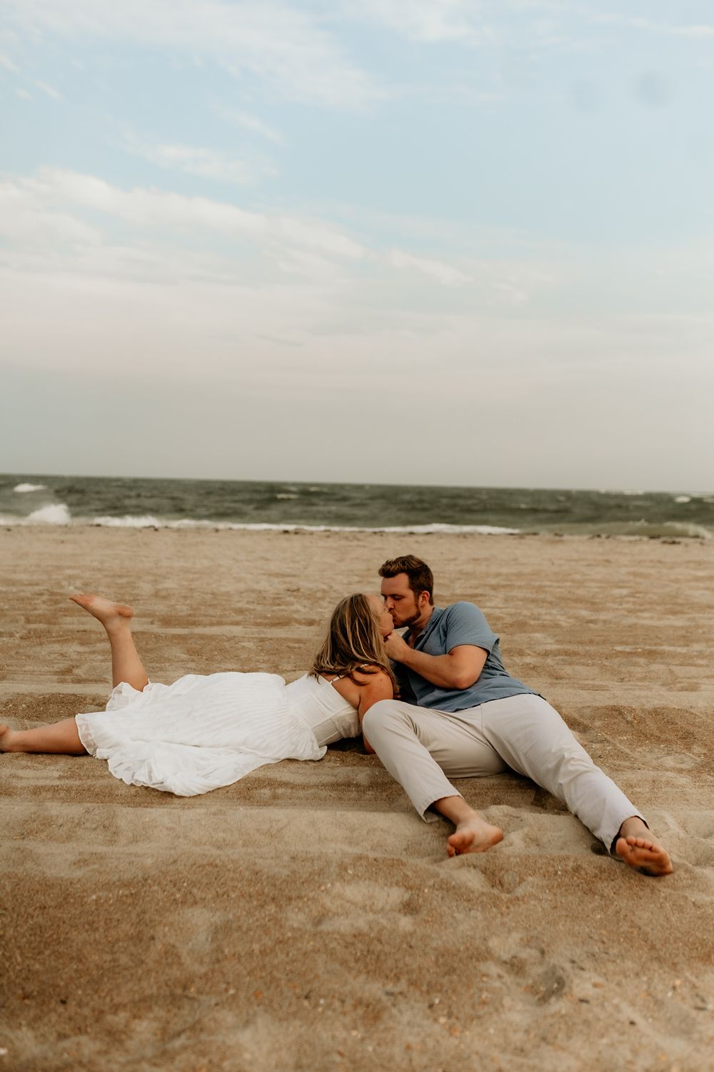 Couple kissing on sandy beach, woman in white dress lying down, man in blue shirt seated, ocean waves in background.