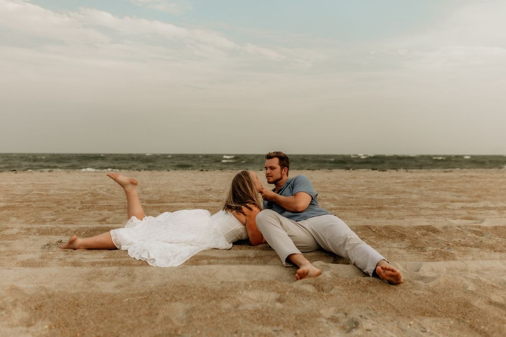Couple relaxing on sandy beach, woman in white dress lying down, man sitting beside her, sharing a romantic moment.