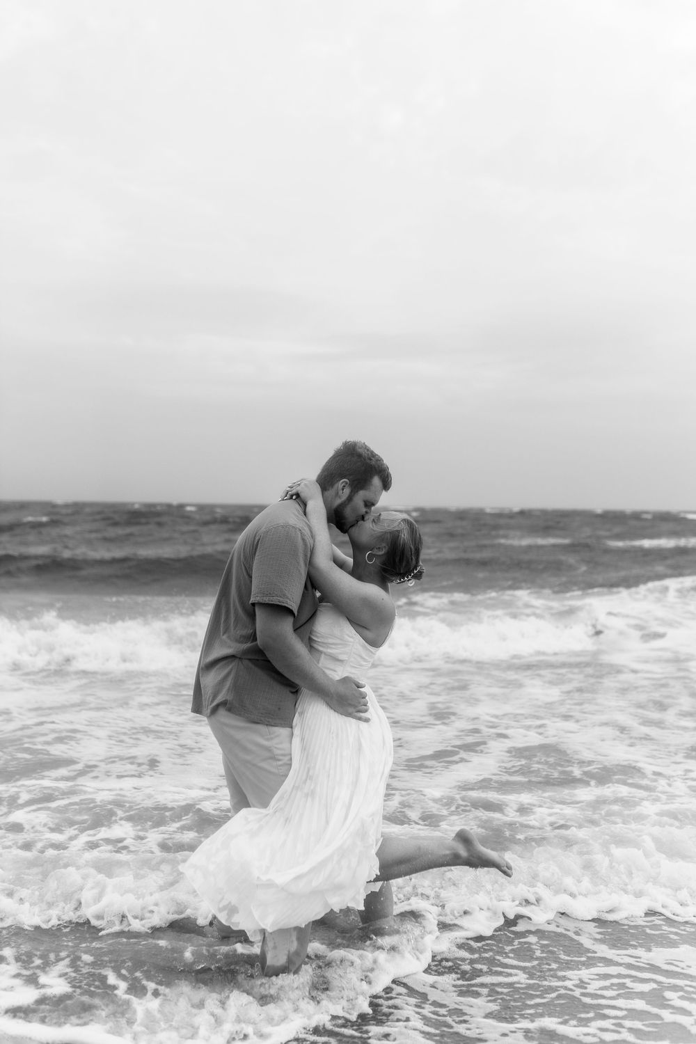 Black and white photo of a couple kissing in ocean waves on a beach, woman in white dress lifted by man.